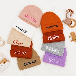 May include: A collection of eight knitted beanies in various colors, each with a different name embroidered on the front. The beanies are arranged in a semi-circle on a white surface.