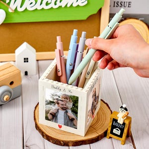 Peut inclure: Un pot à crayons blanc avec une photo d'un père et d'un enfant, et le texte "Daddy & Me". Le pot contient plusieurs stylos aux couleurs pastel. Un petit panneau de boulangerie en bois et d'autres objets décoratifs sont également présents.