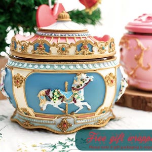 Puede incluir: Una caja de música decorativa con diseño de carrusel en azul, crema y dorado. Presenta un motivo de caballo de carrusel. Otra caja de música rosa es visible en el fondo.