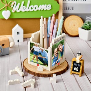Può includere: Un portapenne color crema, progettato per assomigliare a mattoncini, mostra foto di famiglia e la scritta "daddy & Me". Contiene diverse penne dai colori pastello. Il supporto è posto su una fetta di legno, con mattoncini aggiuntivi sparsi.