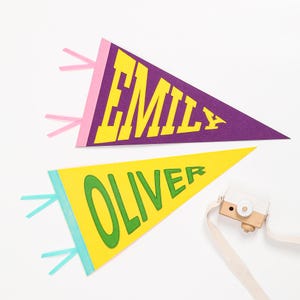 K&ouml;nnte beinhalten: Zwei Filz-Wimpel mit den Namen "Emily" und "Oliver" in bunten Blockbuchstaben. Die Wimpel sind lila und gelb mit rosa und blauen Quasten.