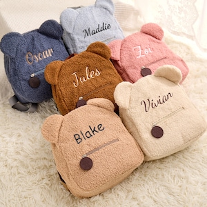 May include: Six plush teddy bear backpacks in various colors, including brown, blue, pink, and cream. Each backpack has a zipper closure and a small pocket on the front. The backpacks are personalized with names, including Oscar, Maddie, Zoi, Jules, Blake, and Vivian.