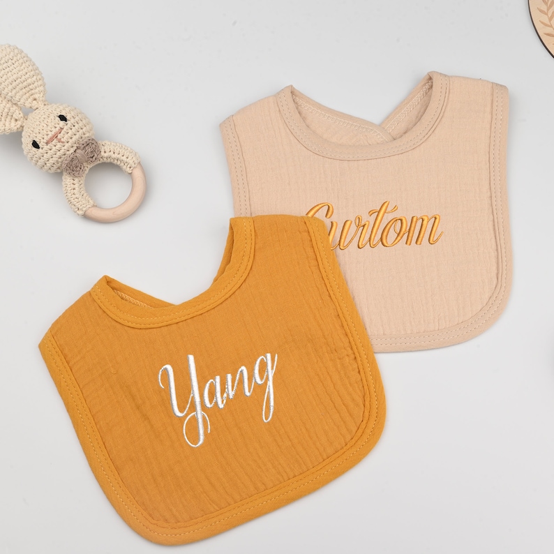 May include: Two baby bibs, one mustard yellow and one beige, with embroidered names "Yang" and "Curtom" in white and gold thread.