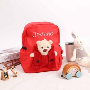 Op de afbeelding: Rode gepersonaliseerde rugzak met een teddybeerzak. De rugzak heeft de naam "Joanne" geborduurd op de voorkant.