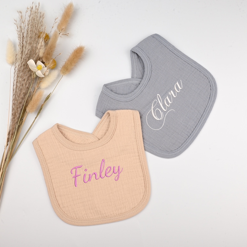 May include: Two personalized baby bibs, one beige with the name "Finley" embroidered in pink and one gray with the name "Clara" embroidered in white.