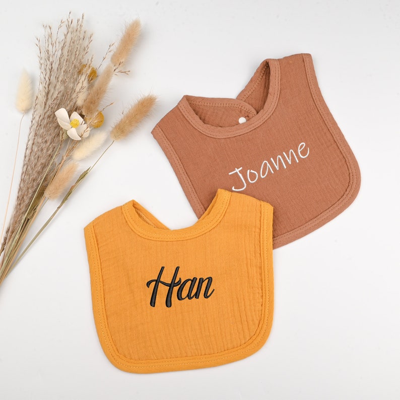 May include: Two personalized baby bibs, one mustard yellow with the name "Han" embroidered in black, and one brown with the name "Joanne" embroidered in white.