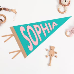 K&ouml;nnte beinhalten: Ein t&uuml;rkisfarbener Filz-Wimpel mit dem Namen "SOPHIA" in rosa Filzbuchstaben. Der Wimpel hat braune Stoffb&auml;nder.