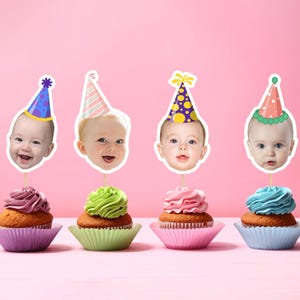 Könnte beinhalten: Vier Cupcakes mit Babygesicht-Toppern, die Partyhüte tragen. Jeder Cupcake hat ein anderes farbiges Frosting: lila, grün, rosa und blau. Die Cupcakes stehen in bunten Papierförmchen vor einem rosa Hintergrund.