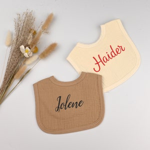 May include: Two personalized baby bibs, one brown with the name "Jolene" embroidered in black and one cream with the name "Haider" embroidered in red.