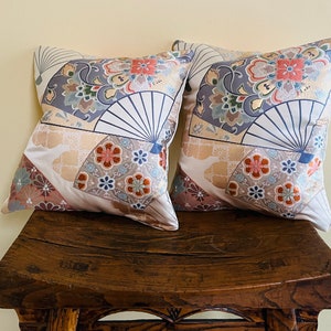 May include: Two decorative throw pillows with a Japanese-inspired design featuring fans, flowers, and geometric patterns in shades of blue, gold, and pink. The pillows are sitting on a wooden bench.