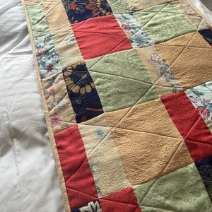 May include: A quilted table runner featuring a patchwork design. The runner incorporates various fabrics in shades of red, blue, green, and beige, with floral and geometric patterns. The edges are finished with a beige border.
