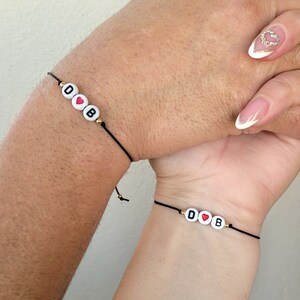 Könnte beinhalten: Zwei schwarze Armbänder aus Schnur mit weißen Perlen, die an jedem Handgelenk "D♥B" buchstabieren.