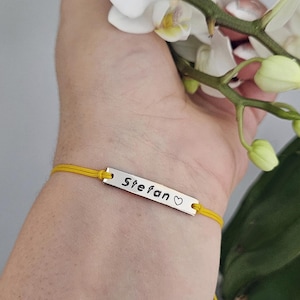 Op de afbeelding: Een geel koordarmband met een rechthoekige zilveren bedel. De bedel is gegraveerd met de naam "Stefan" en een hartsymbool. De armband wordt om een pols gedragen, met witte bloemen op de achtergrond.