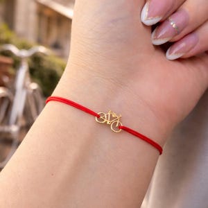 May include: A red string bracelet featuring a small gold-colored bicycle charm. The bracelet is simple and delicate, with the bicycle charm as the focal point. The bracelet is worn on a wrist.