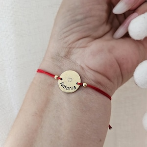 Può includere: Un braccialetto con un disco dorato con il nome "Antonia" e un cuore. Il disco è attaccato a un cordino rosso con perline dorate. Il braccialetto è indossato al polso.