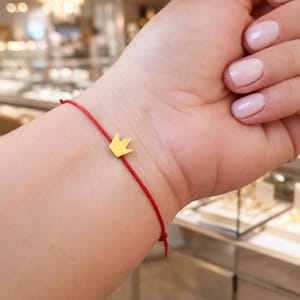May include: A red string bracelet featuring a small gold crown charm. The crown is positioned centrally on the bracelet. The bracelet is secured with a knot. The background is out of focus.