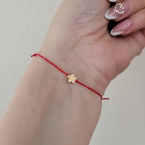 May include: A red string bracelet with a small, gold-colored flower charm. The bracelet is tied around a wrist, showcasing the delicate design and vibrant color. The flower charm is centered on the string.