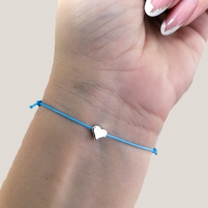 May include: A light blue string bracelet with a small, silver heart charm. The bracelet is tied on the wrist. The heart charm is centered on the string.