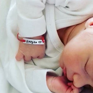 Puede incluir: Un bebé durmiendo con un body blanco. Una pulsera roja y blanca con el nombre "Sergiu" y una corona está en la muñeca del bebé. La cara del bebé es visible, con los ojos cerrados y una mano cerca de la boca.
