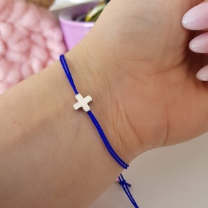 May include: A blue string bracelet with a silver cross charm. The bracelet is tied with a knot and has two silver beads at the end.