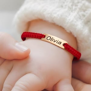 以下が含まれることがあります： 「Olivia」と書かれた金色のネームプレートが付いた赤い織りブレスレットのクローズアップ。ブレスレットは赤ちゃんの腕にあり、大人の手で持たれています。赤ちゃんは白いセーターを着ています。