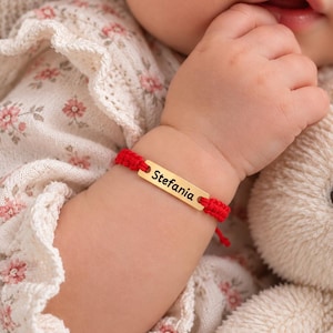 May include: A red cord bracelet featuring a rectangular gold-toned metal bar engraved with the name Stefania in black script. The bracelet is adjustable with knotted cord ends and is worn on a wrist next to a plush toy and floral fabric.