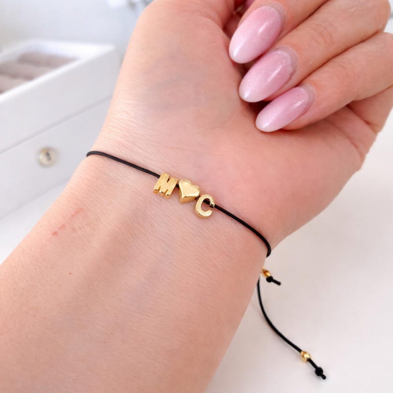 May include: A black string bracelet with gold-colored letter beads spelling "M&hearts;C". The bracelet is on a wrist with pink nail polish. The bracelet has adjustable sliding knots.