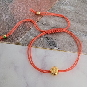 May include: Coral-colored adjustable bracelet featuring a gold-colored Buddha head charm. The bracelet is made of a thin, woven cord with gold-colored beads at the ends. The Buddha head charm is detailed with facial features.