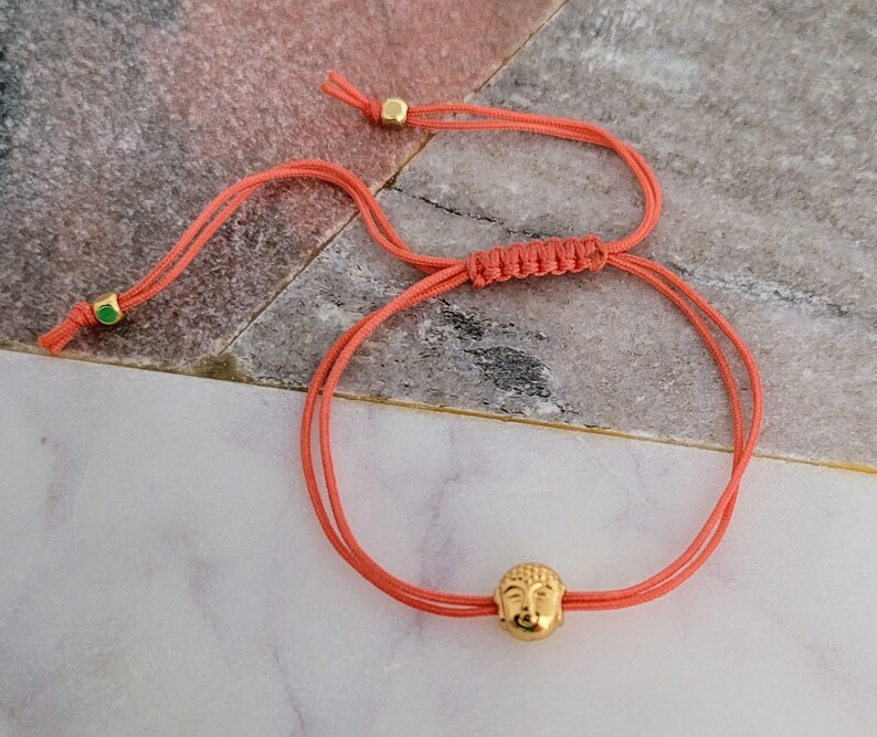 May include: Coral-colored adjustable bracelet featuring a gold-colored Buddha head charm. The bracelet is made of a thin, woven cord with gold-colored beads at the ends. The Buddha head charm is detailed with facial features.