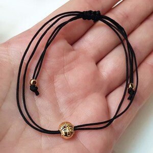 May include: Black cord bracelet featuring a gold-colored Buddha head charm. The bracelet has adjustable sliding knots and small gold beads. The Buddha head is intricately detailed, and the bracelet is designed to be worn on the wrist.