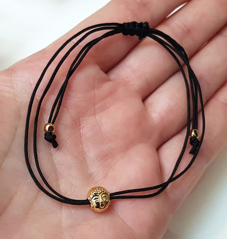 May include: Black cord bracelet featuring a gold-colored Buddha head charm. The bracelet has adjustable sliding knots and small gold beads. The Buddha head is intricately detailed, and the bracelet is designed to be worn on the wrist.