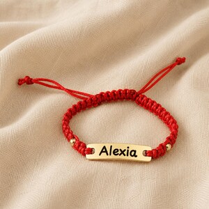 Könnte beinhalten: Ein rotes geflochtenes Armband mit einer mittigen, goldfarbenen Metallplatte, auf der der Name Alexia in schwarzer Schrift eingraviert ist. Das Armband enthält zwei kleine goldfarbene Perlen neben der Platte und verstellbare Knotenenden.