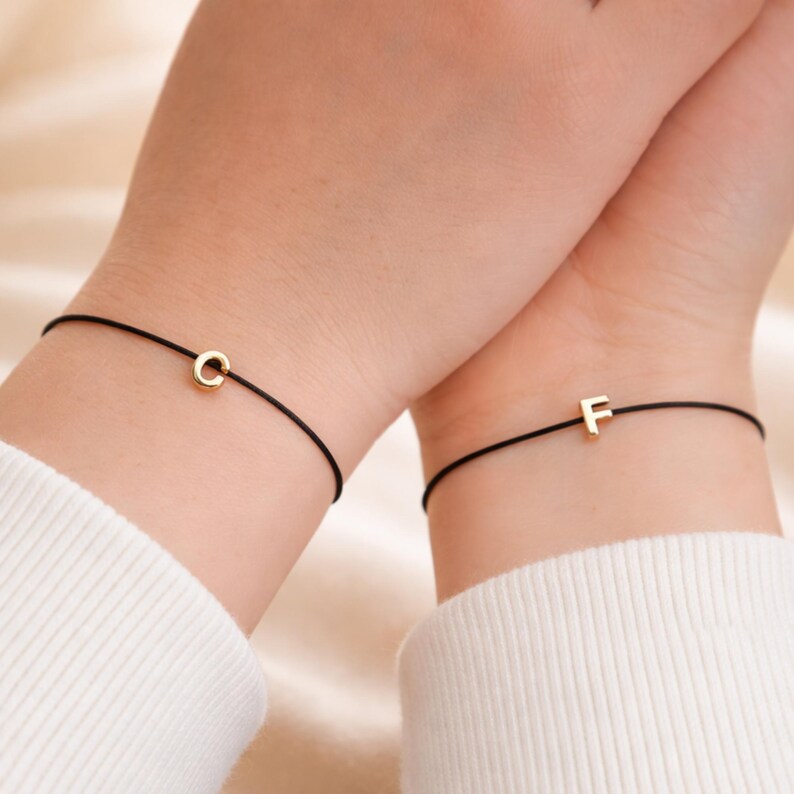 May include: Two black string bracelets, each with a small gold initial charm. One bracelet features the letter "C", and the other displays the letter "F". The bracelets are worn on wrists, with white sleeves visible in the background.