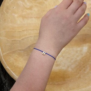 May include: A gold-colored letter "B" charm bracelet on a blue cord. The bracelet is simple and delicate, with the charm centered on the cord. The bracelet is worn on a wrist.