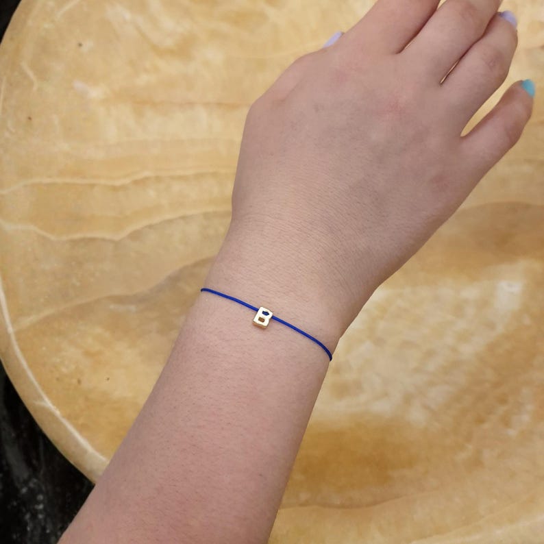 May include: A gold-colored letter "B" charm bracelet on a blue cord. The bracelet is simple and delicate, with the charm centered on the cord. The bracelet is worn on a wrist.