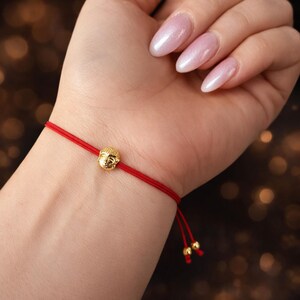 May include: A red string bracelet featuring a small, gold-colored Buddha head charm. The bracelet is tied with a sliding knot for adjustable sizing. The Buddha charm is detailed with facial features.