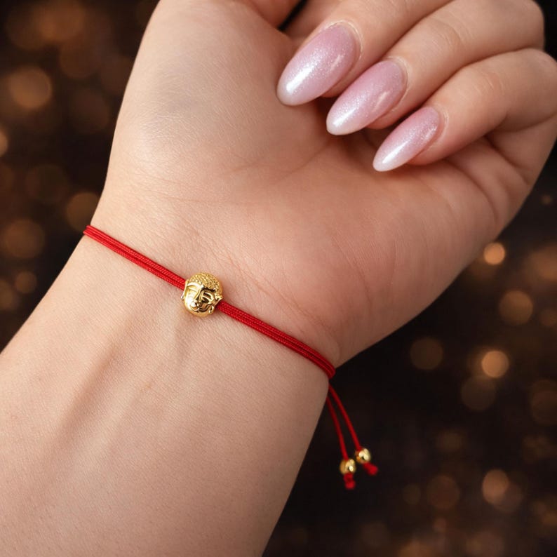 May include: A red string bracelet featuring a small, gold-colored Buddha head charm. The bracelet is tied with a sliding knot for adjustable sizing. The Buddha charm is detailed with facial features.