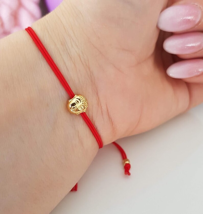 May include: A red string bracelet with a gold-colored charm. The charm is round with a detailed design. The bracelet is tied around a wrist.