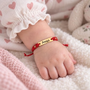 Puede incluir: Una pulsera de cordón rojo con una placa rectangular de metal dorado grabada con el nombre Anays. La pulsera incluye pequeñas cuentas doradas integradas en el diseño de cordón rojo trenzado, ajustable con nudos corredizos.