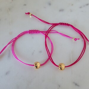 May include: Two adjustable bracelets with bright pink cords and gold-colored Buddha head charms. The bracelets have a sliding knot closure and small gold beads at the ends. The background is a light-colored, possibly marble, surface.