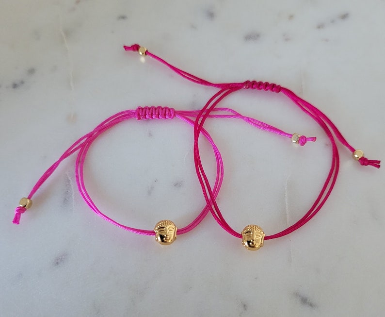 May include: Two adjustable bracelets with bright pink cords and gold-colored Buddha head charms. The bracelets have a sliding knot closure and small gold beads at the ends. The background is a light-colored, possibly marble, surface.