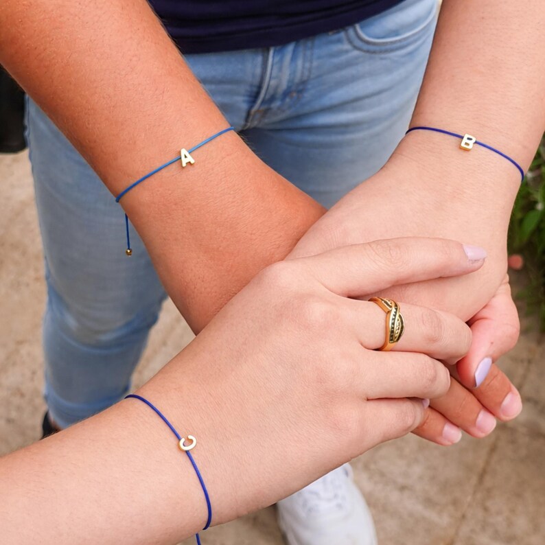 May include: Three blue string bracelets, each with a gold initial: A, B, and C. The bracelets are worn on wrists, with a gold ring on a finger. The background includes blue jeans and a navy blue shirt.
