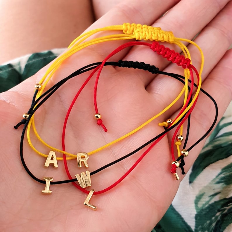 May include: A collection of colorful friendship bracelets. The bracelets are made of yellow, red, and black cords, with gold-colored letter beads spelling "APRIL". Each bracelet has an adjustable knot closure and small gold beads.