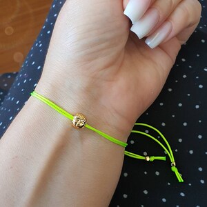 May include: A neon green string bracelet featuring a small gold-colored Buddha head charm. The bracelet is tied with adjustable knots. The bracelet is shown on a wrist.