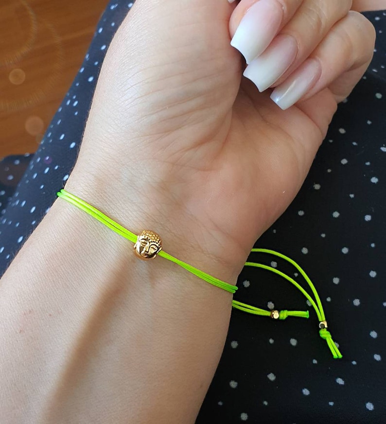 May include: A neon green string bracelet featuring a small gold-colored Buddha head charm. The bracelet is tied with adjustable knots. The bracelet is shown on a wrist.