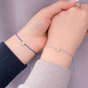 May include: Two matching bracelets, one with a gold letter "N" on a blue cord and the other with a gold letter "A" on a light blue cord. The bracelets are worn on wrists, showcasing a minimalist design.