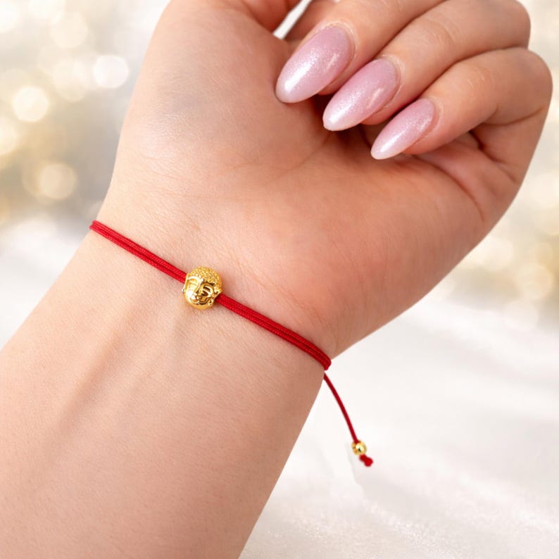May include: A red string bracelet featuring a small, gold-colored Buddha head charm. The bracelet is tied around a wrist, showcasing the simple yet elegant design. The background is softly blurred, highlighting the bracelet.