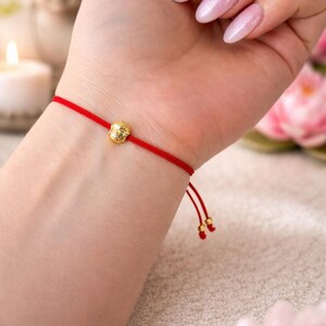 May include: Red string bracelet with a gold-colored Buddha charm. The bracelet is tied with a sliding knot for adjustable sizing. The Buddha charm is a small, detailed sculpture. The bracelet is a simple, elegant accessory.