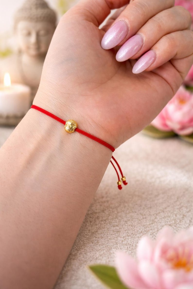 May include: Red string bracelet with a gold-colored Buddha charm. The bracelet is tied with a sliding knot for adjustable sizing. The Buddha charm is a small, detailed sculpture. The bracelet is a simple, elegant accessory.