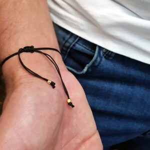 May include: Black adjustable bracelet with gold-colored beads. The bracelet is tied with a sliding knot closure. The bracelet is worn on a wrist, with a white shirt and blue jeans in the background.